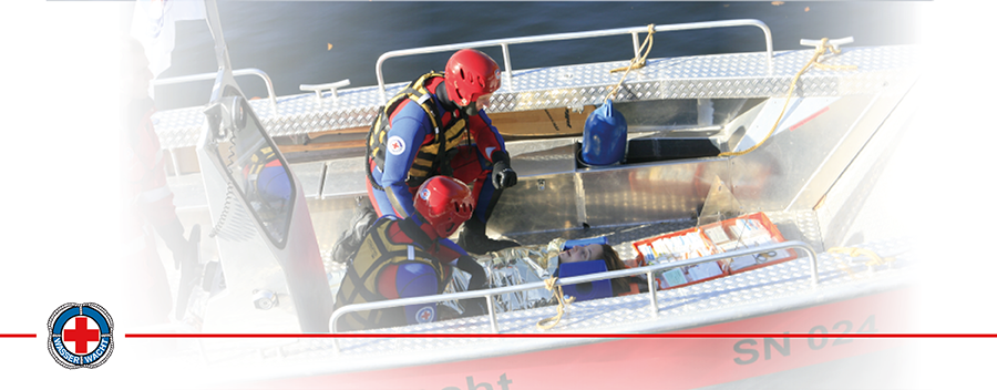 Zwei Wasserretter knien auf einem Boot und behandeln eine verletzte Person, die auf dem Boden des Bootes liegt.