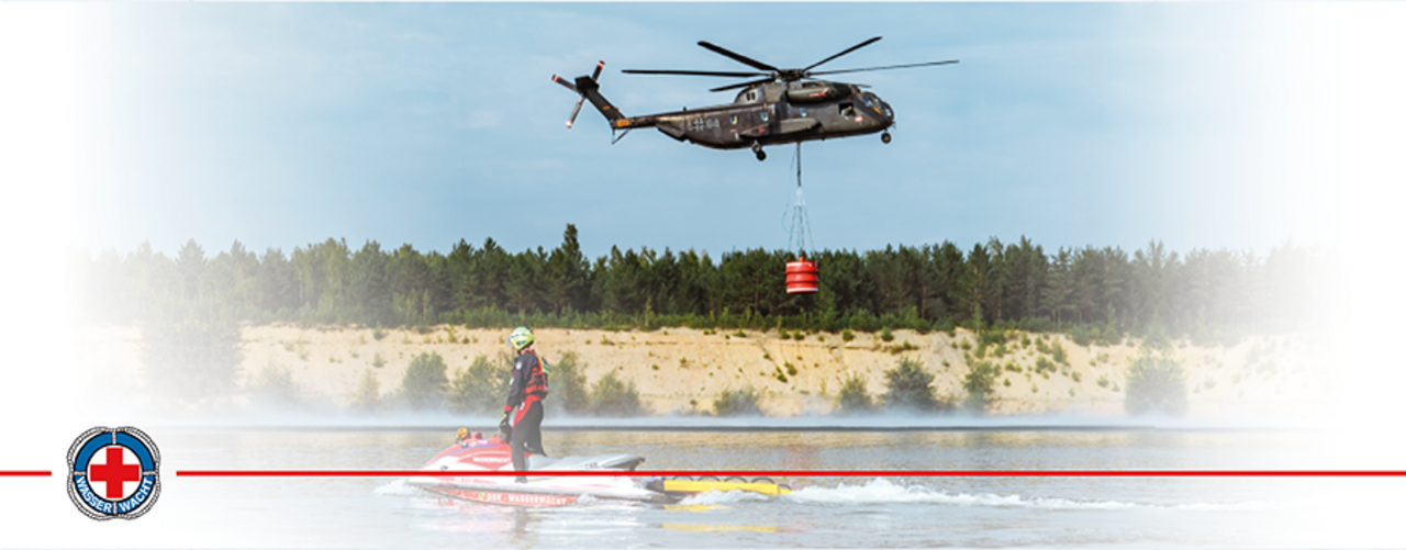 Ein Hubschrauber fliegt über einem See. Unter ihm steht ein Ehrenamtlicher der Wasserwacht auf seinem Wasserfahrzeug und schaut nach oben.